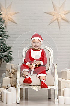 Happy little smiling boy in santa claus costume sitting on armchair near christmas tree and holding christmas gift box