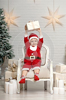 Happy little smiling boy in santa claus costume sitting on armchair near christmas tree and holding christmas gift box