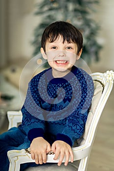 Happy little smiling boy sitting on armchair near christmas tree and holding christmas gift box