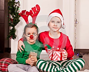 Happy little smiling girl and boy in Santa cap and deer mask sitting on the blanket in Christmas pajamas with gift box.