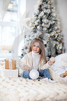 Happy little smiling girl with gift on Christmas Eve sit on bed. Child opens New year gift near christmas tree with glowing lights