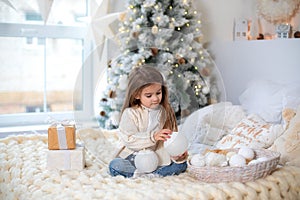Happy little smiling girl with gift on Christmas Eve sit on bed. Child opens New year gift near christmas tree with glowing lights
