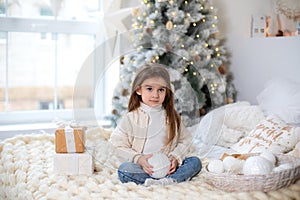 Happy little smiling girl with gift on Christmas Eve sit on bed. Child opens New year gift near christmas tree with glowing lights