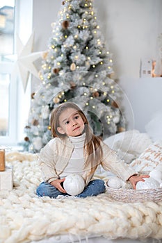 Happy little smiling girl with gift on Christmas Eve sit on bed. Child opens New year gift near christmas tree with glowing lights
