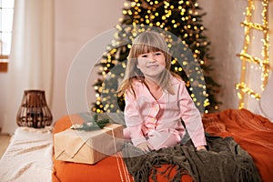 Happy little smiling girl in pajamas with gift on xmas Eve lies on bed. child opens New year gift at home near christmas tree with