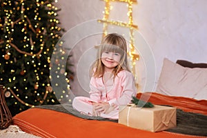 Happy little smiling girl in pajamas with gift on xmas Eve lies on bed. child opens New year gift at home near christmas tree