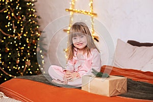 Happy little smiling girl in pajamas with gift on xmas Eve lies on bed. child opens New year gift at home near christmas tree with