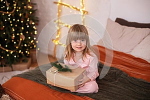 Happy little smiling girl in pajamas with gift on xmas Eve lies on bed. child opens New year gift at home near christmas tree with