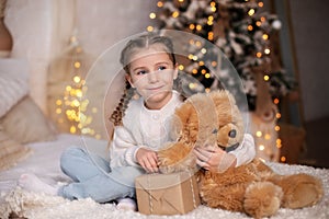 Happy little smiling girl with pigtails opens christmas gift at home. Child in a white sweater lies on bed on Christmas Eve and wi