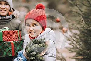 Happy little smiling girl in red cap and boy with Christmas gift boxes standing near by spruce. Holiday celebrate concept