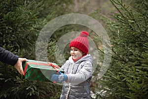 Happy little smiling girl in red cap takes a given box with a gift for Christmas. Kid standing near by spruce. Holiday celebrate