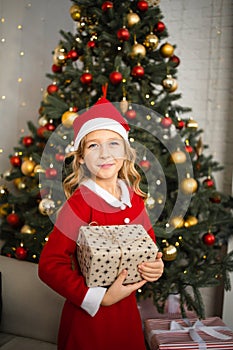 Happy little smiling girl in santa hat with christmas gift box stanging near Christmas tree.