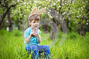 Happy little toddler boy eating chocolate and wearing Easter bunny ears, sitting in blooming garden on warm sunny day. Celebrating