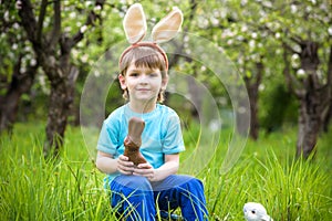 Happy little toddler boy eating chocolate and wearing Easter bunny ears, sitting in blooming garden on warm sunny day. Celebrating