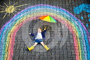 Happy little toddler girl in rubber boots with rainbow sun and clouds with rain painted with colorful chalks on ground