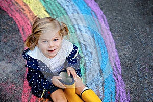 Happy little toddler girl in rubber boots with rainbow sun and clouds with rain painted with colorful chalks on ground