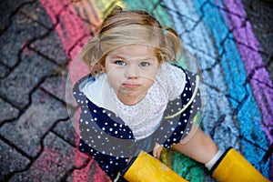 Happy little toddler girl in rubber boots with rainbow sun and clouds with rain painted with colorful chalks on ground