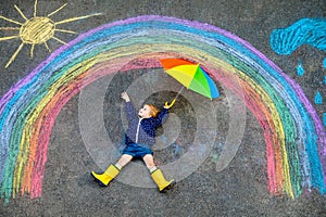 Happy little toddler girl in rubber boots with rainbow sun and clouds with rain painted with colorful chalks on ground