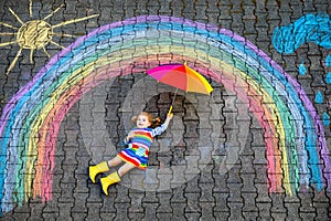 Happy little toddler girl in rubber boots with rainbow sun and clouds with rain painted with colorful chalks on ground