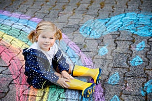 Happy little toddler girl in rubber boots with rainbow sun and clouds with rain painted with colorful chalks on ground