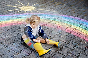 Happy little toddler girl in rubber boots with rainbow sun and clouds with rain painted with colorful chalks on ground
