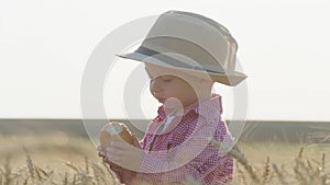 Happy little toddler in hat eats bread while standing in wheat field at sunset. Summer country life and agriculture