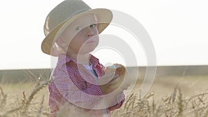 Happy little toddler in hat eats bread while standing in wheat field at sunset. Summer country life and agriculture