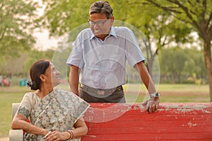 Happy looking retired senior Indian man and woman couple, sitting on a red bench, smiling and looking at each other in a park