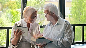 Happy lovely retired couple standing opposite each other with tablet and book. They smile.