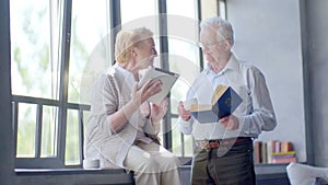 Happy lovely retired couple standing opposite each other with tablet and book. Smile and drink tea