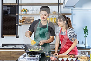 Happy lovely young Asian couple with casual clothes is smiling, helping for preparing and cooking food together in modern kitchen