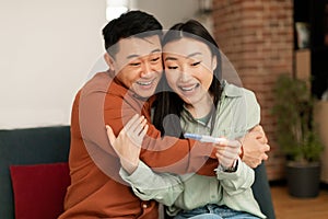 Happy loving asian family learning about their future child from pregnancy test, man embracing wife, sitting on sofa