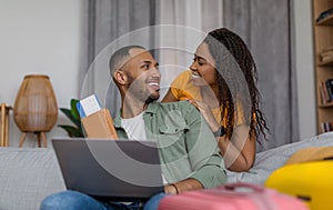 Happy loving black spouses ready for travel, holding boarding pass and using laptop, planning vacation trip together