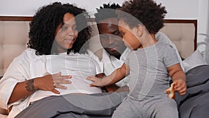 Happy loving couple admiring son touching pregnant belly sitting on bed with parents. Smiling African American mother