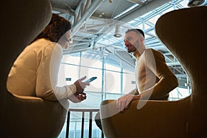 Happy loving couple, business partners, handsome man and pretty woman resting together, sitting on armchairs in airport departure