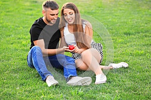 Happy loving couple with red heart sitting on green grass outdoors