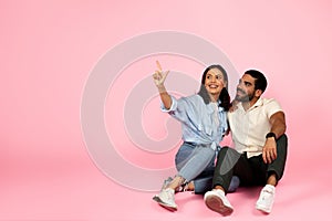 Happy loving couple sitting on floor, pointing at copy space