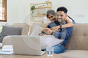 Happy loving family Indian couple hugging on couch at home and focused on laptop screen watching romantic movie