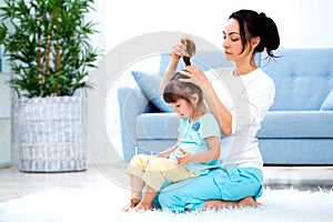 Happy loving family. Mother is combing her daughter`s hair sitting on the carpet on the floor in the room