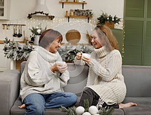 Happy loving family mother and grown up daughter sitting in kitchen decorated on Christmas enjoying hot tea and talking to each