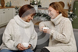 Happy loving family mother and grown up daughter sitting in kitchen decorated on Christmas enjoying hot tea and talking to each