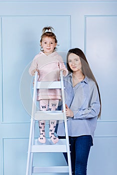 Happy loving family. young Mother and her little daughter standing on blue ladder playing and hugging on blue wall background.