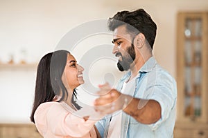 Happy loving indian couple dancing waltz, holding hands, having fun together at home on weekend, side view