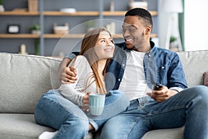 Happy loving interracial couple relaxing at home, watching tv and drinking coffee