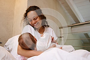 Happy loving mother smiles while connects with her baby, holds him in her arms, sitting on the bed in white bedchamber