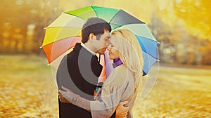 Happy loving young couple kissing and hugging under colorful umbrella in autumn park on warm sunny day