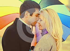 Happy loving young couple kissing and hugging under colorful umbrella in autumn park on warm sunny day