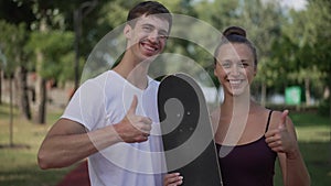 Happy loving young couple with skateboard looking at each other turning to camera gesturing thumbs up smiling. Portrait