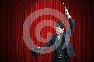 Happy magician or illusionist is showing magic trick with his wand. Red curtains in background