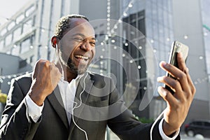 Happy male African American businessman standing outside office center wearing headphones and talking to his team